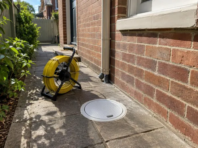 Flush capped inspection opening beside a coiled drain camera reel on a concrete side path by a brick house in Randwick