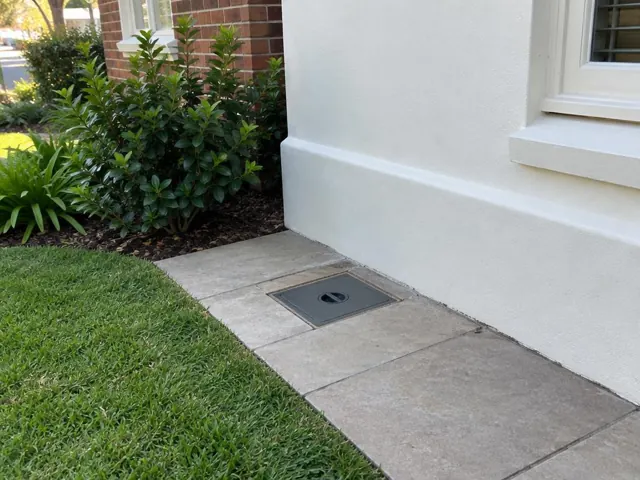 Flush capped access cover in paved side path beside a house wall and garden in Coogee