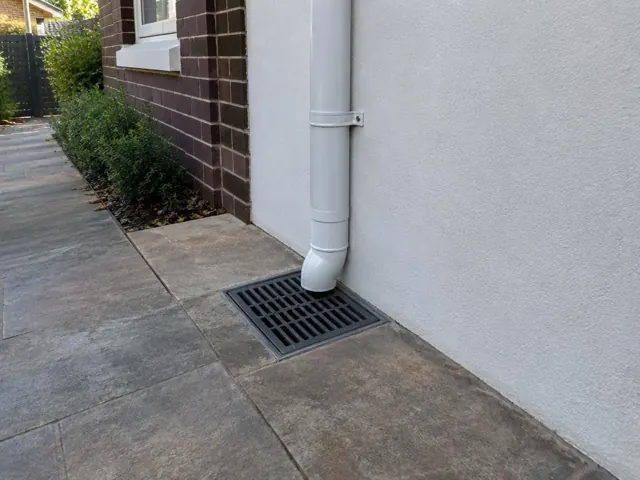 Stormwater grate beside a downpipe on a paved side path next to a brick and rendered wall in Kensington